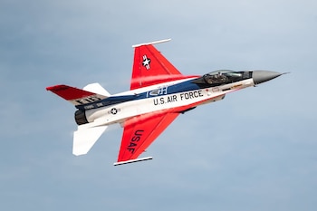 Avión de combate F-16 Viper de la Fuerza Aérea de EE. UU. en vuelo ascendente contra un cielo azul claro. La aeronave es roja, blanca y azul, con estelas de vapor y el piloto visible
