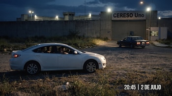Una vista nocturna del CERESO Uno con un muro alto, torre de vigilancia y entrada principal. Un automóvil blanco con personas y otro oscuro salen.