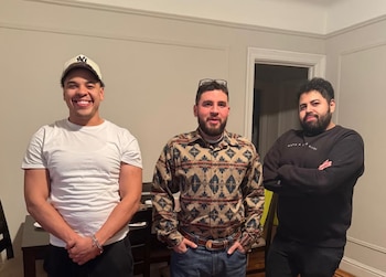 Tres hombres jóvenes posan de pie en el interior de una habitación frente a una mesa y una pared blanca