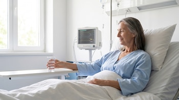 Mujer adulta mayor con cabello gris en bata azul, sentada en cama de hospital blanca, sostiene vaso de agua. Monitor médico y gotero de suero visibles al fondo.