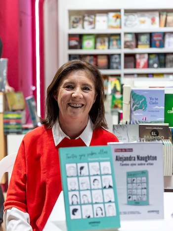 Retrato de la escritora Alejandra Naughton, una mujer de mediana edad con cabello castaño y suéter rojo, sonriendo frente a un stand con sus libros y estanterías