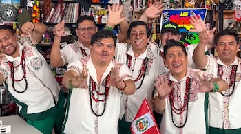 Siete integrantes del grupo Los Mirlos sonríen y saludan con las manos en alto, vestidos con camisas blancas bordadas y collares.
