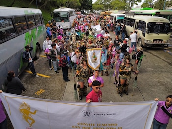Multitud de personas con trajes de colores y tocados; un autobús verde, dos blancos, uno beige; pancarta blanca con texto y logos; calle asfaltada