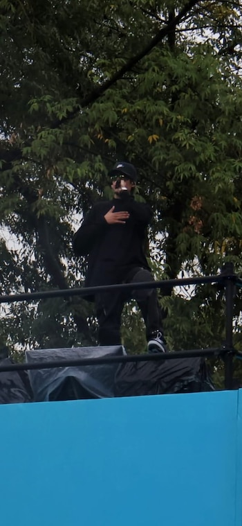 Hombre con gorra, gafas de sol, ropa negra, zapatillas blancas y micrófono en escenario con barandales negros y un fondo de árboles