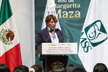 Mujer de cabello castaño corto, vestida de azul, habla en un podio con micrófono. Detrás, banderas de México y del IMSS y un cartel con texto