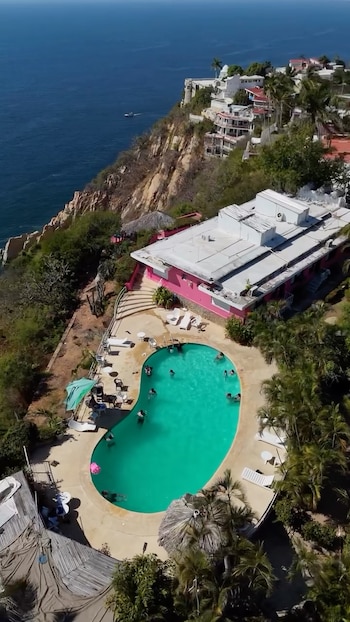 Vista aérea de una piscina con personas nadando, junto a un edificio rosado en un acantilado frente al mar