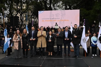 Un grupo de hombres y mujeres de pie en un escenario, algunos aplaudiendo, y niños con banderas. Detrás, una pantalla con la frase 'La ciudad homenajea al Padre de la Patria'