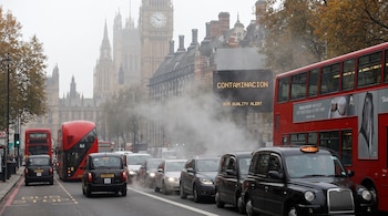 Calle de Londres con tráfico denso de taxis y autobuses rojos, humo de escape, señal digital con 'CONTAMINACION', edificios históricos y árboles.