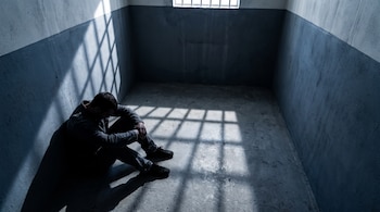 Hombre sentado con la cabeza baja en una celda vacía con la sombra de barrotes proyectada en el suelo y la pared.
