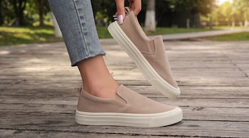 Persona con pantalón de mezclilla y una zapatilla slip-on beige, sostiene otra zapatilla igual sobre un sendero de madera con árboles de fondo.
