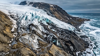Vista aérea de un glaciar con grietas, terreno rocoso oscuro y bloques de hielo flotando en la costa marina.