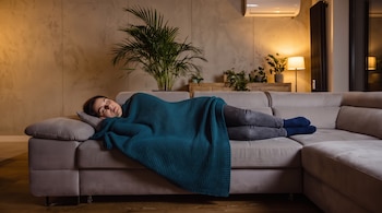 Una mujer duerme de lado en un sofá gris con una manta azul verdoso. Se observa un cojín, pantalones, calcetines, un aire acondicionado, plantas y una lámpara.