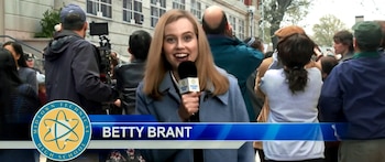 Una mujer con cabello rubio sostiene un micrófono en una mano, sonriendo a cámara. Un logo de "Midtown Technical High School" y "Betty Brant" aparecen en pantalla
