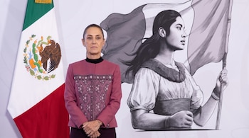 Mujer de pie con traje rosa oscuro y cabello recogido, frente a la bandera de México y una ilustración a escala de una mujer que sostiene una bandera.