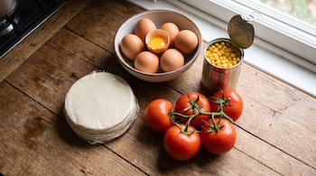 Una pila de tapas para empanadas, un cuenco con huevos, uno roto con la yema visible, una lata abierta de choclo en grano y tomates en rama sobre una superficie de madera.