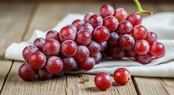 Racimo de uvas rojas recién lavadas con pequeñas gotas de agua visibles en su piel, sobre un paño blanco y una tabla de madera.