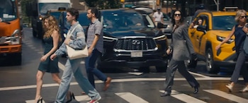 Vista de calle con peatones cruzando un paso de cebra en una ciudad. Una mujer con traje gris y gafas de sol en primer plano, flanqueada por otras personas y vehículos