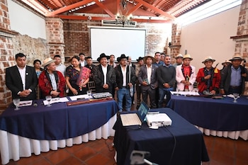 Grupo de personas, mayoritariamente hombres, de pie detrás de mesas con tela azul. Hay documentos, micrófonos, vasos, laptop, proyector y pantalla blanca