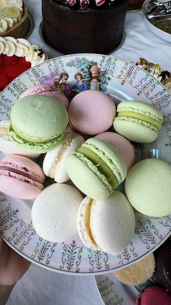 Un plato de porcelana con macarons verdes, rosados y blancos, rodeado de tartas y un pastel de chocolate sobre un mantel blanco