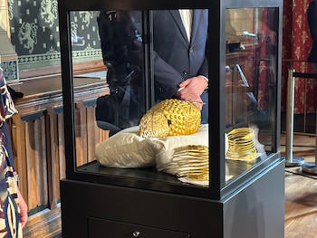 El Casco de Coțofenești, un casco de oro con relieves, se exhibe en una vitrina transparente sobre un cojín blanco. Se ven brazaletes dorados y el reflejo de una persona en traje