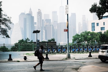 Una persona camina frente al horizonte del Bajo Manhattan mientras una densa capa de neblina y contaminación cubre parte del skyline de Nueva York, fenómeno asociado a las altas temperaturas y la mala calidad del aire en la ciudad.