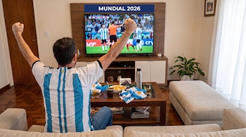 Hombre con camiseta de Argentina y brazos en alto frente a un televisor que muestra un partido de fútbol con el texto Mundial 2026, mate y papas fritas en una mesa.