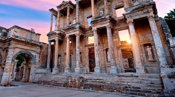 Gran edificio de piedra con columnas, escaleras de varios niveles y un arco, visto al atardecer bajo un cielo con tonos rosas y azules.