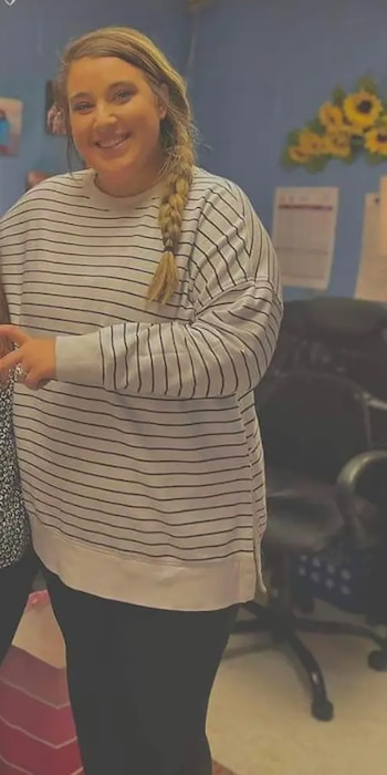 Una mujer de cabello rubio trenzado sonríe a la cámara, vestida con una camiseta de rayas horizontales blancas y negras, con un fondo azul