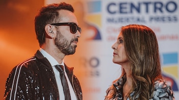 Primer plano de un hombre con gafas de sol y una mujer, ambos de perfil, mirándose fijamente con un fondo borroso de escenario.