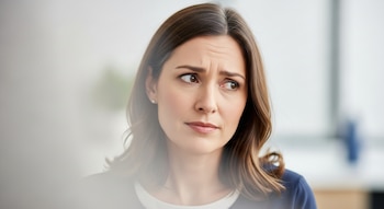 Retrato de una mujer de mediana edad, con cabello castaño y canas, el ceño fruncido y la mirada perdida. Viste ropa oscura sobre una prenda blanca.