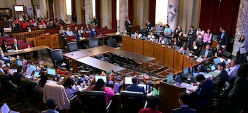 Vista aérea de una sala grande con columnas y paneles de madera donde se celebra una conferencia. Personas sentadas en escritorios con banderas y laptops, y audiencia en gradas