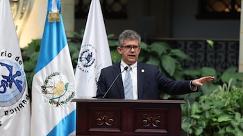 Un hombre de mediana edad con gafas y traje oscuro habla desde un podio de madera. Detrás hay banderas de Guatemala y plantas