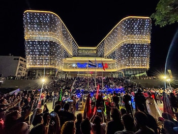 Vista nocturna de un edificio moderno con luces horizontales en forma de 'V', una multitud observando un concierto con músicos y personas disfrazadas en primer plano