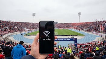 Primer plano de una mano sosteniendo un smartphone con el mensaje 'SIN SEÑAL' en pantalla, y un estadio de fútbol con gradas llenas de gente al fondo.