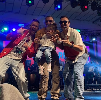 Niña con gafas rosas sostenida por un hombre en un escenario. Otro hombre con trompeta a la izquierda, y uno con gafas de sol a la derecha. Luces de colores de fondo