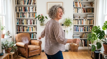Mujer de 60 años con cabello gris y rizado sonriendo y bailando en un salón con estanterías llenas de libros, plantas y un sillón de cuero.