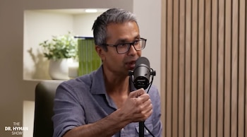 Hombre de cabello gris y gafas habla en un micrófono negro en un estudio. Se ven estantes con plantas y una pared de madera a rayas detrás