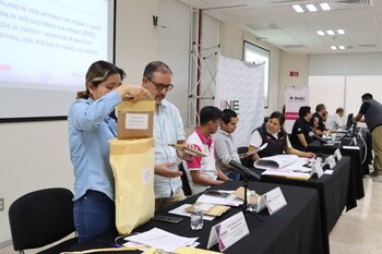 En un evento del INE, una mujer y un hombre manipulan sobres de votación. Se ven otras personas en mesas y un proyector con información sobre el voto electrónico