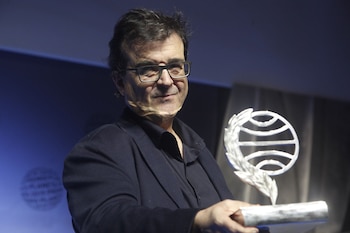Un hombre con gafas, camisa y chaqueta oscuras sostiene un trofeo metálico en forma de globo terráqueo con una rama de laurel