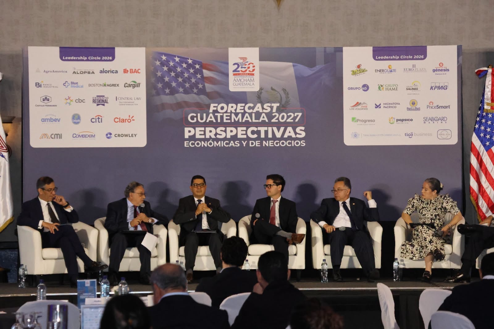 Un panel de discusión en el evento Forecast Guatemala 2027 aborda perspectivas económicas y de negocios, con la bandera de Estados Unidos y Guatemala al fondo. (AmchAm Guatemala)