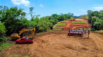 Excavadora amarilla, camioneta roja, camino de tierra. Obras de construcción vial con terraplenes y bosque. Cartel de madera con texto Anillo Regional C-50.