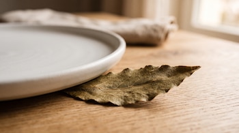 Una hoja de laurel seca situada bajo el borde de un plato blanco de cerámica sobre una mesa de madera con una servilleta al fondo.
