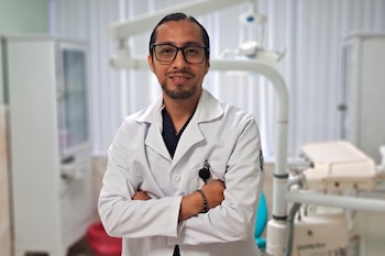 Un hombre con barba y gafas, vestido con una bata blanca sobre un uniforme oscuro, está de pie con los brazos cruzados en un consultorio médico