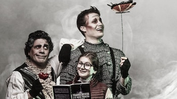 Dos hombres con vestuario teatral y maquillaje junto a una niña con anteojos que sostiene un libro abierto ante un fondo gris con humo