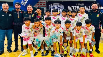 Once niños con ojos borrosos y tres adultos posan en cancha deportiva. Los niños visten uniformes, medallas y llevan bolsas con objetos. Un trofeo y balón en el suelo.