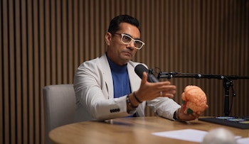 Un hombre con gafas y chaqueta clara sostiene un modelo de cerebro naranja mientras habla ante un micrófono en un estudio con fondo de madera acanalada