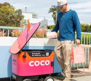 Un hombre de gorra y camisa azul recibe un paquete de un robot de reparto Coco de color rosa y naranja con la tapa abierta, en un entorno exterior