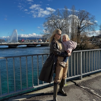 Una mujer sostiene en brazos a una niña junto a una barandilla metálica, con un lago, un puente y un chorro de agua de fondo