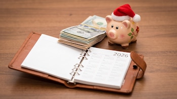 Una agenda abierta con un calendario de 2026, un fajo de billetes de cien dólares y una alcancía de cerdo con gorro navideño sobre madera.