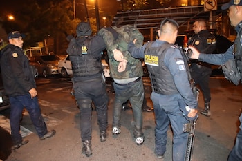Cuatro agentes de policía con chalecos antibalas y uniformes oscuros detienen a un hombre esposado vestido de camuflaje en una calle oscura por la noche